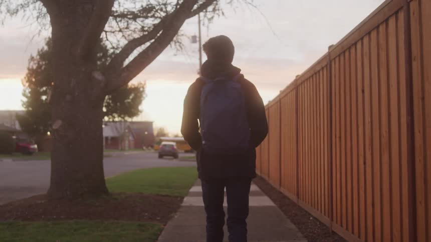 The video shows a person walking away from the camera down a residential sidewalk at dusk they are wearing a dark jacket jeans and a blue backpack and are walking alongside a wooden fence and past a large tree