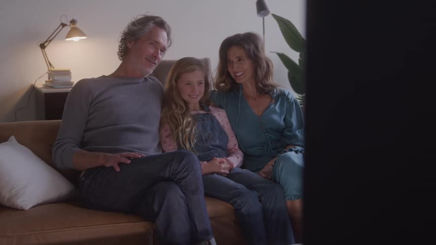 A family of three is sitting on a brown couch and watching tv together the parents are on either side of their young daughter and they are all smiling