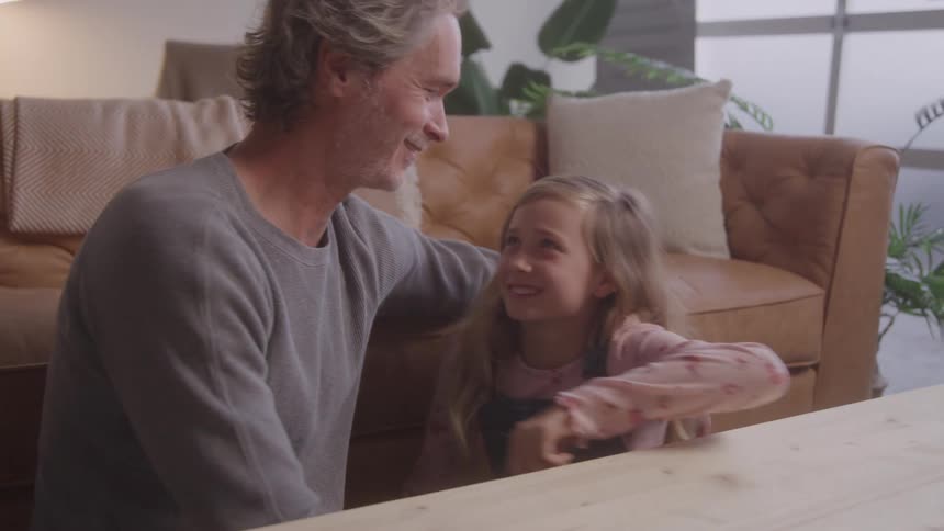 Father and daughter sitting on the floor of the living room with the sofa in the background embrace each other affectionately