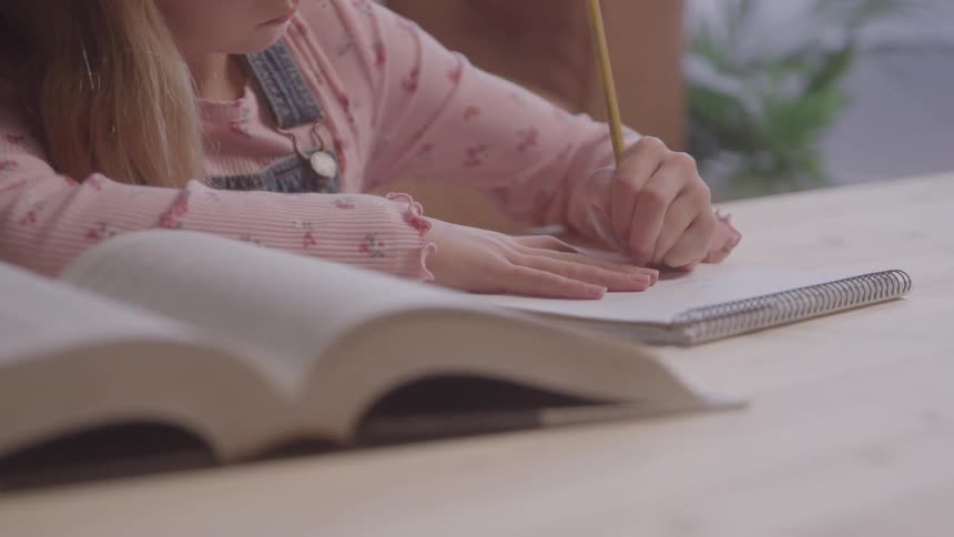 A female student wearing a pink long sleeved shirt and overalls sits at a table and writes in a notebook with a pencil with a book open in front of her