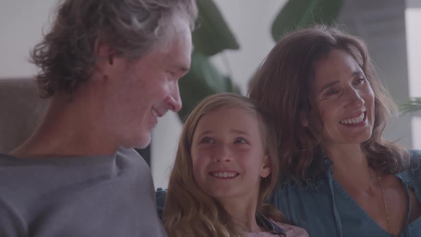 A teenage girl sits between her parents as they all smile at each other then look forward afterward indoors at midday