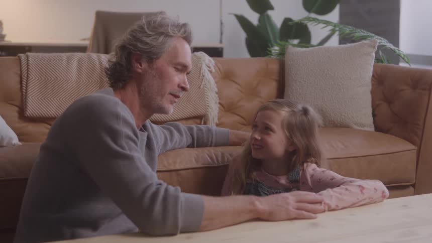 A man and a young girl sit by a wooden table smiling warmly at each other in a cozy living room