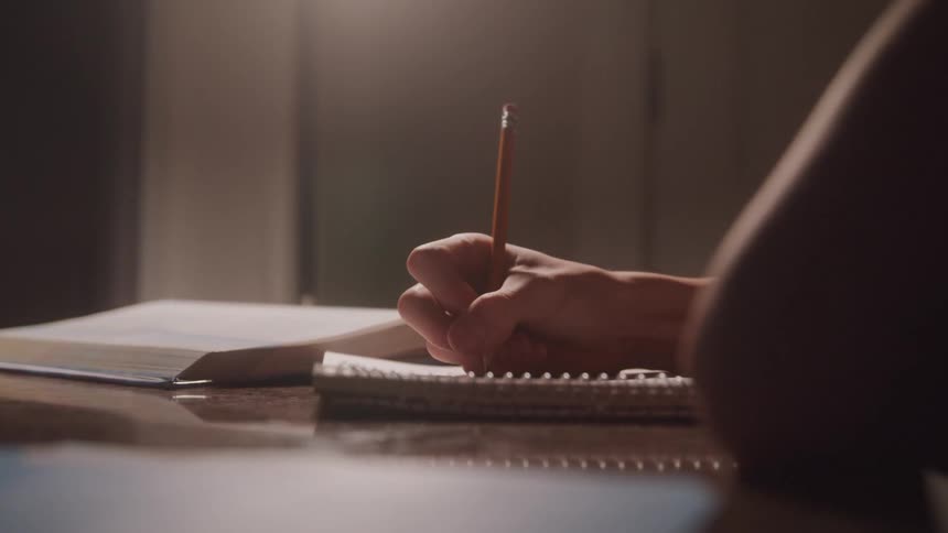 A person writes in a notebook with a pencil in soft natural light