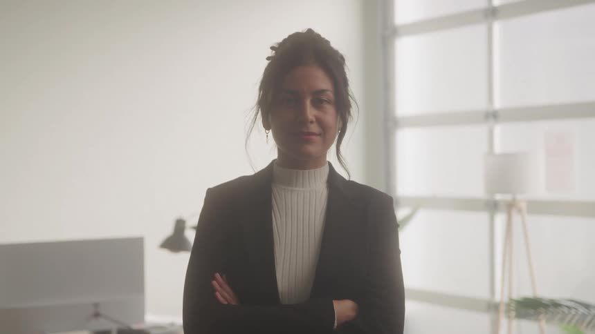 A professionallooking woman with her hair in a bun stands confidently in an office setting she is wearing a black blazer over a white turtleneck and has her arms crossed