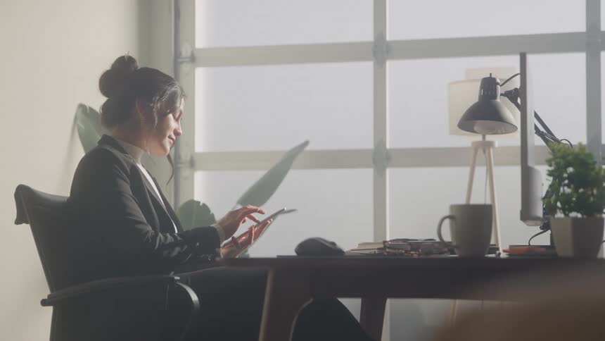 A young woman with her hair in a bun is sitting at a desk in a bright office working on a tablet she is wearing a black blazer and is partially silhouetted against large windows in the background