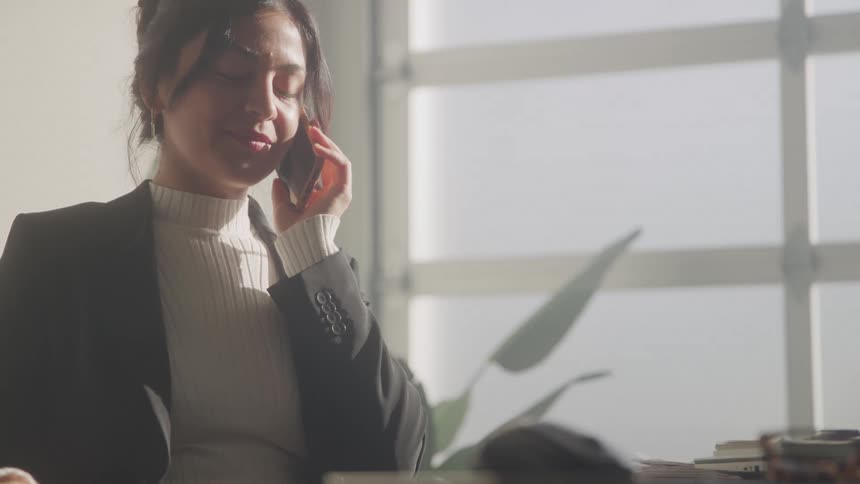 A smiling professional woman with her hair pulled back wears a black blazer over a creamcolored turtleneck while talking on a smartphone next to a sunlit window