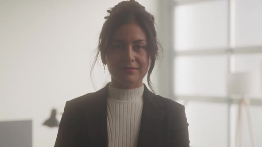 A young woman wearing a turtleneck shirt a suit and earrings stands inside an office at midday with a smile on her face