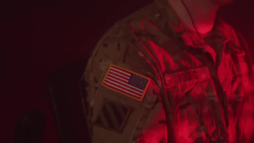 A closeup of a persons torso wearing a military camouflage uniform with an american flag patch on the sleeve the scene is dimly lit with a red light