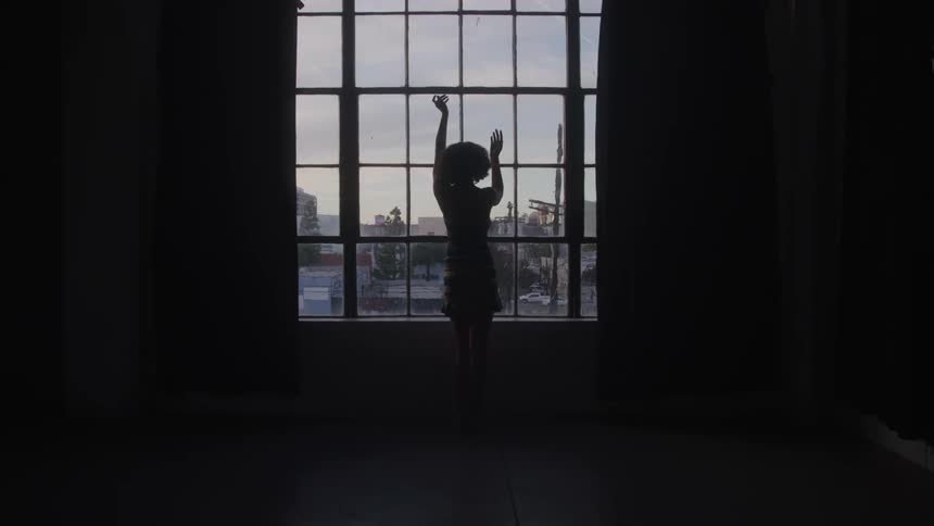 A silhouette of a dancer with an afro is seen from behind standing in front of a large window with multiple panes the person stretches their arms upwards as if in a dance pose