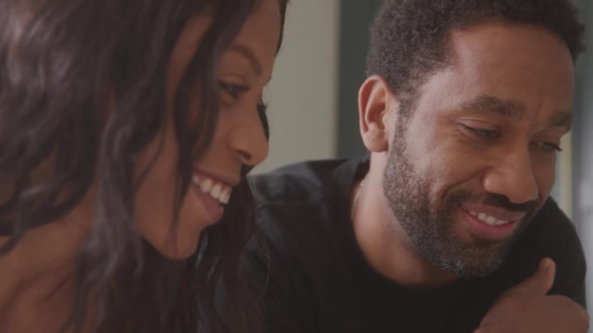 A closeup of a smiling woman with long dark hair and a man with a beard sitting together the woman is laughing while looking at the man who is looking down and smiling