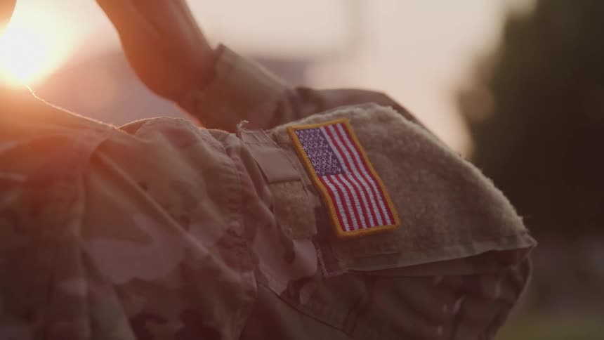 A military person is shown from the back wearing a camouflage uniform with an american flag patch on the sleeve their hand is seen moving towards their head as if to salute while the sun shines brightly in the background