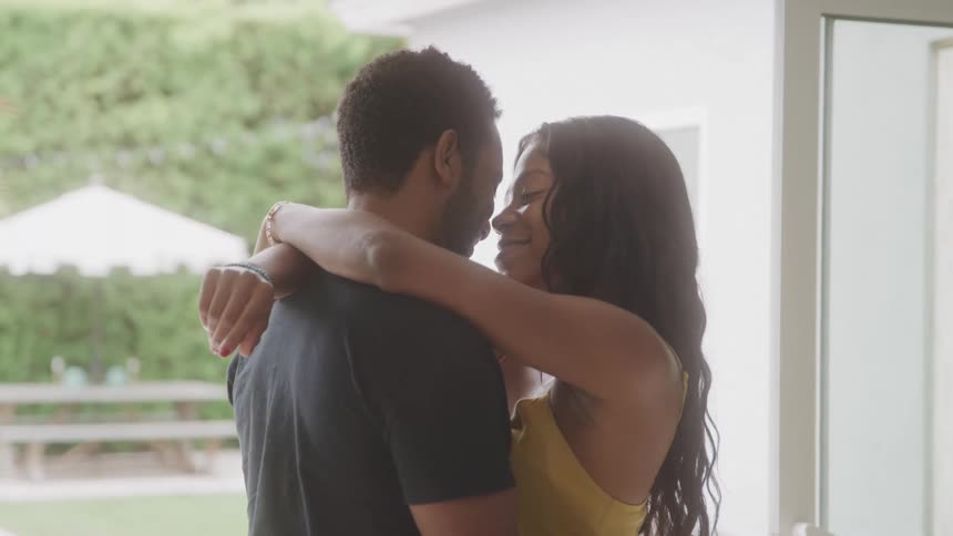 A man and a woman hug each other smiling as they stand beside a glass window at midday