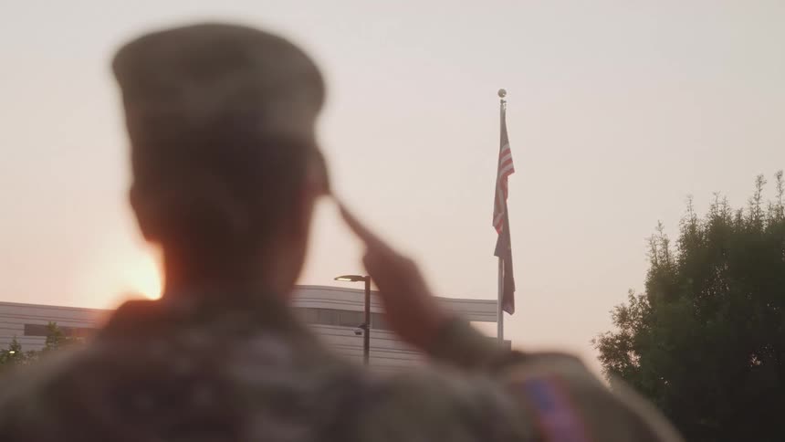 Female soldier standing outdoors salutes the american flag while the sun shines over a nearby building