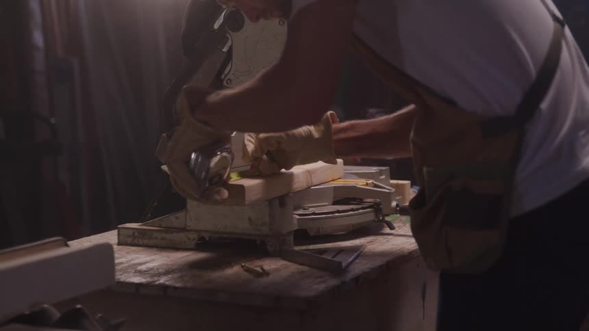 A craftsman wearing an apron and work gloves prepares a miter saw to cut a piece of wood in a dimly lit workshop