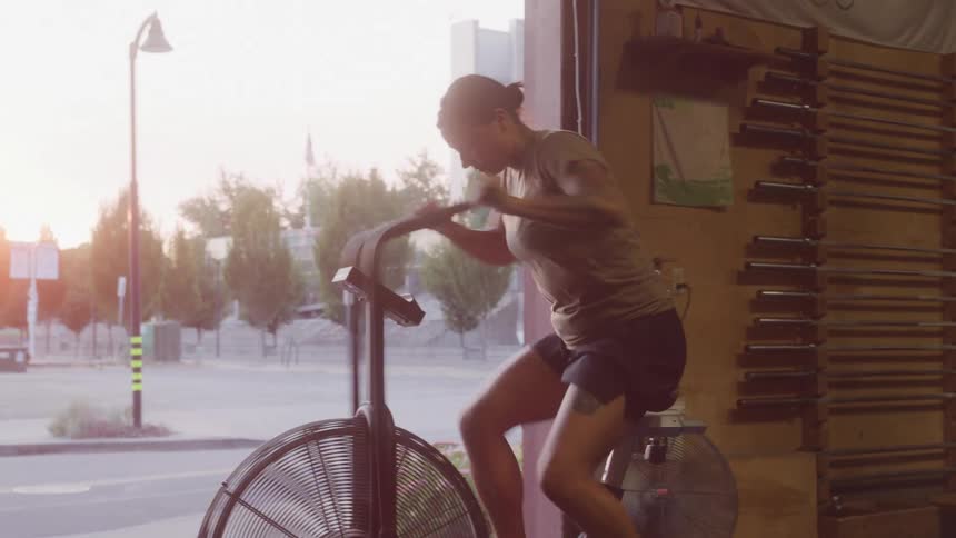 Woman working out intensely on an air bike inside a gym at sunset