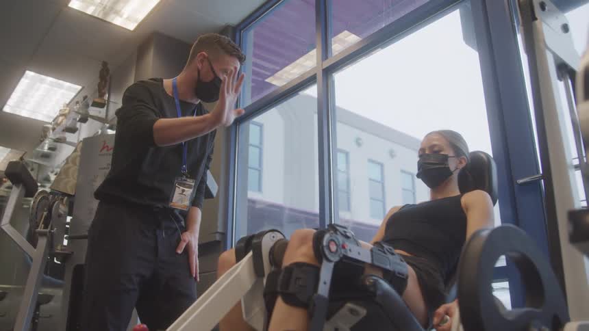A woman in a face mask is lying on a leg press machine with a physical therapist standing beside her the therapist also wearing a mask is holding his hand up as if to give a highfive