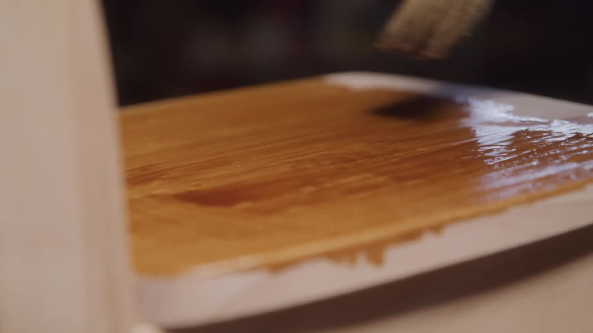 A hand holding a paintbrush applies a clear or lightcolored liquid like varnish or stain to a wooden surface the liquid changes the color of the wood to a richer darker tone