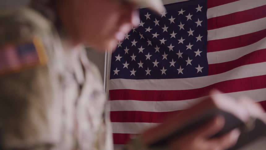 Woman in military uniform reading a bible indoors with the american flag in the background