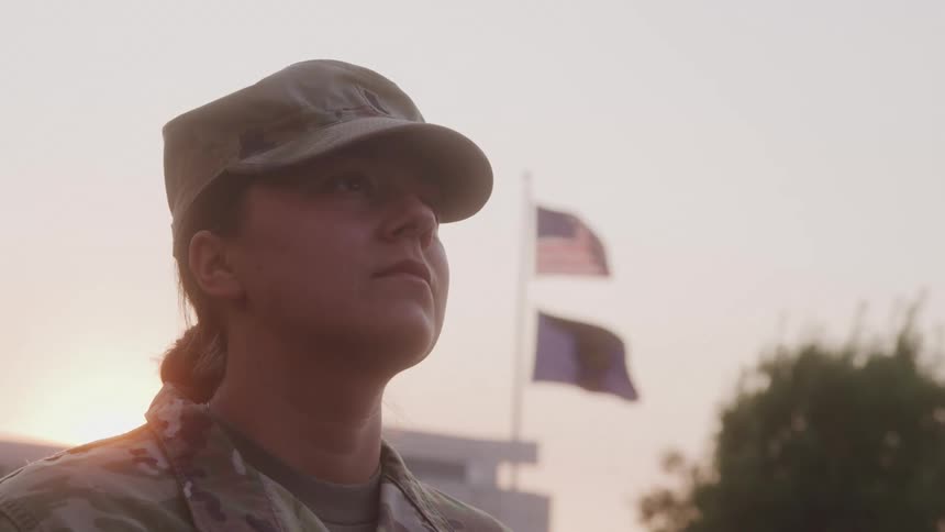 A closeup profile shot of a seriouslooking military woman in uniform with a blurred american flag and military base in the background at dusk