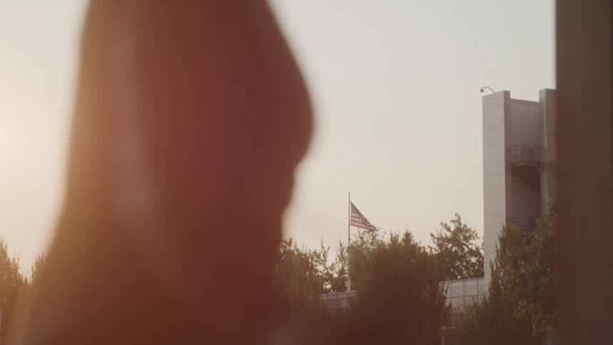 A persons head and shoulder jumping rope silhouetted in the foreground as an american flag trees and a modern building stand in the background under a hazy sky