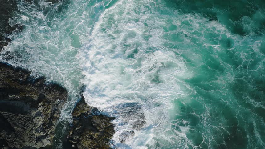 A closeup aerial view shows waves with white foam crashing onto a rocky coastline the water is a vibrant mix of turquoise and deep blue