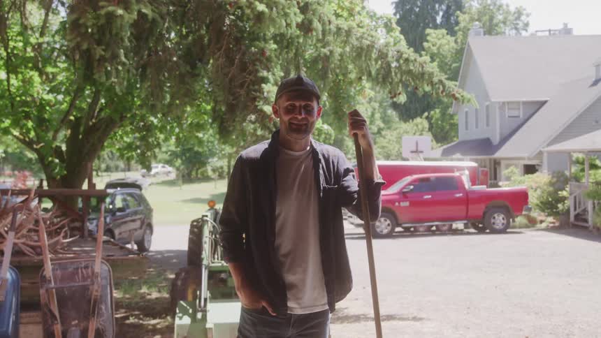 A farmer wearing a cap and a black buttondown shirt stands in front of a red pickup truck and a tractor they are holding a longhandled tool possibly a hoe or rake and smiling at the camera