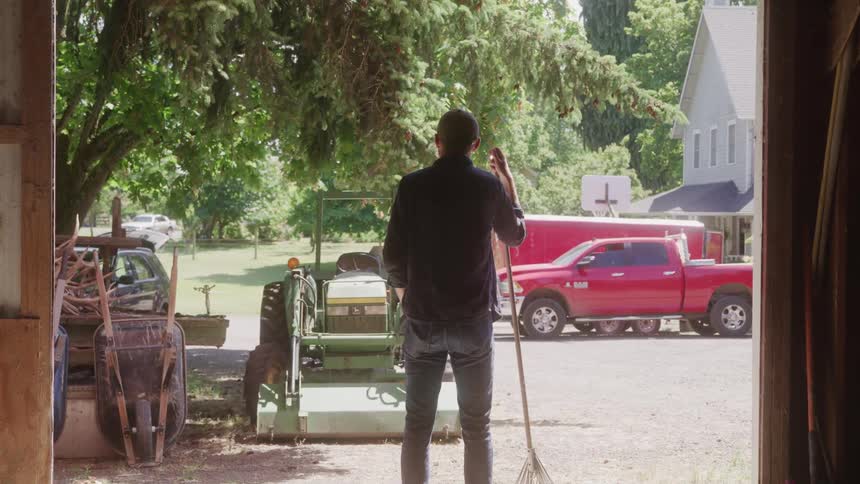 A person is seen from the back standing outside of a barn or garage holding a rake a red pickup truck and a tractor are parked behind them and a house is visible in the background