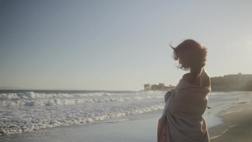The woman is standing on the beach wrapped in a blanket