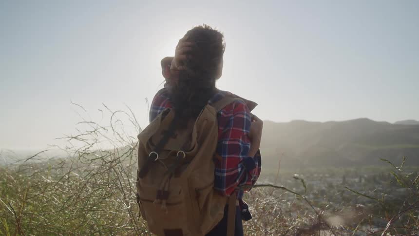 A woman on a hike carrying a backpack stands in a tall grassy area under the sun staring forward