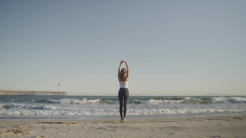 A woman stands on the beach and stretches as waves wash ashore