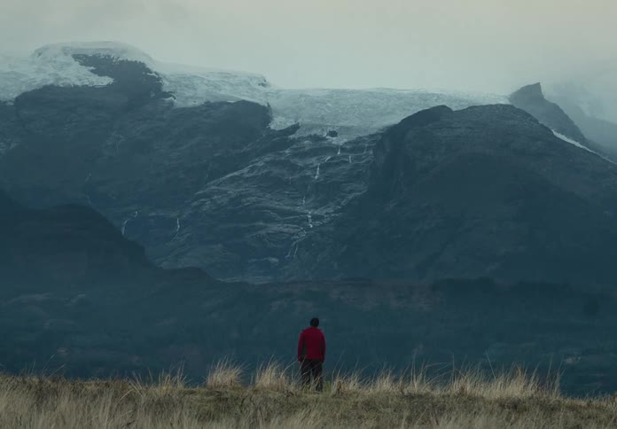 A solitary figure stands in a field of dry grass facing a vast dark mountain range the mountains are shrouded in mist giving the scene a mysterious and somber tone the person is wearing a bright red hoodie which provides a stark contrast against the