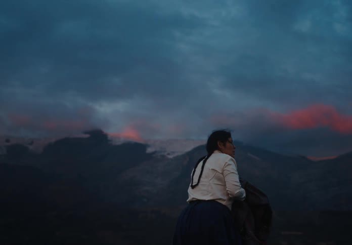 A woman with braided hair stands alone facing a mountain landscape under a moody twilight sky