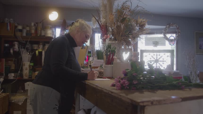 An elderly woman writes on a document with a pen inside a flower shop at midday