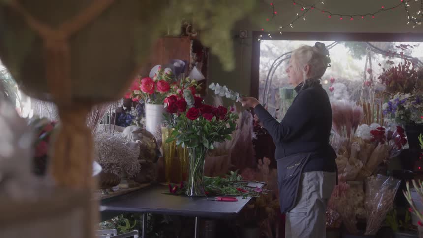 A woman wearing a black longsleeved shirt and a black apron is standing near a work table in a cluttered flower shop arranging a bouquet of red roses and other colorful flowers in a glass vase