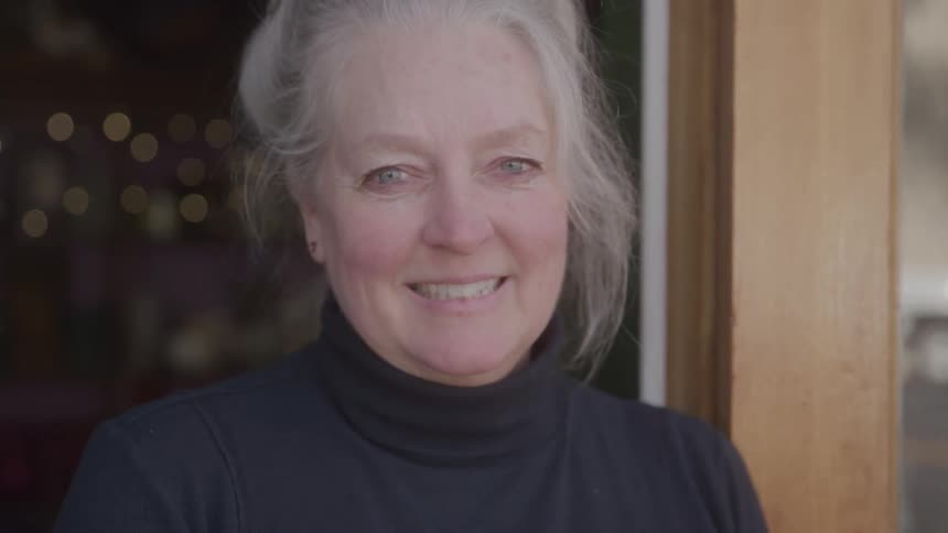 An elderly woman stands on her front porch wearing a black dress and smiling at midday
