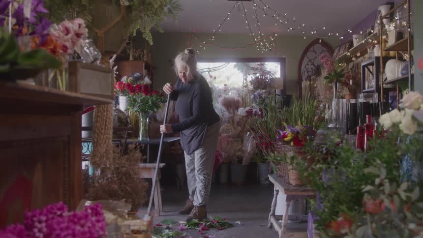 A female florist with blonde hair in a bun wears a black shirt light gray pants and a black apron as she sweeps colorful petals and leaves from the floor of a flower shop with a longhandled broom