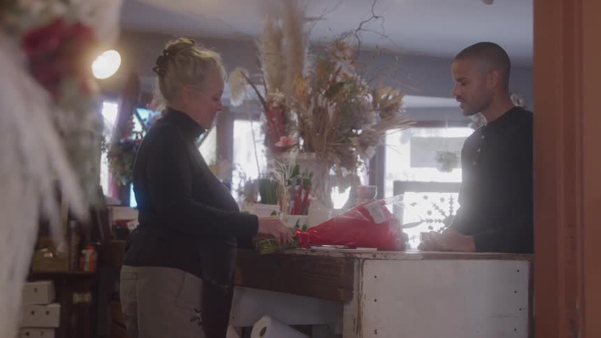 Woman hands a red bouquet to man at a counter surrounded by dried flowers