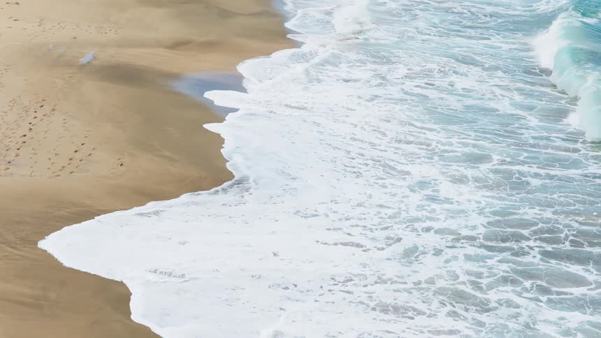 A foamy turquoise ocean wave crashes against a sandy beach leaving a wet line where the water recedes back into the sea