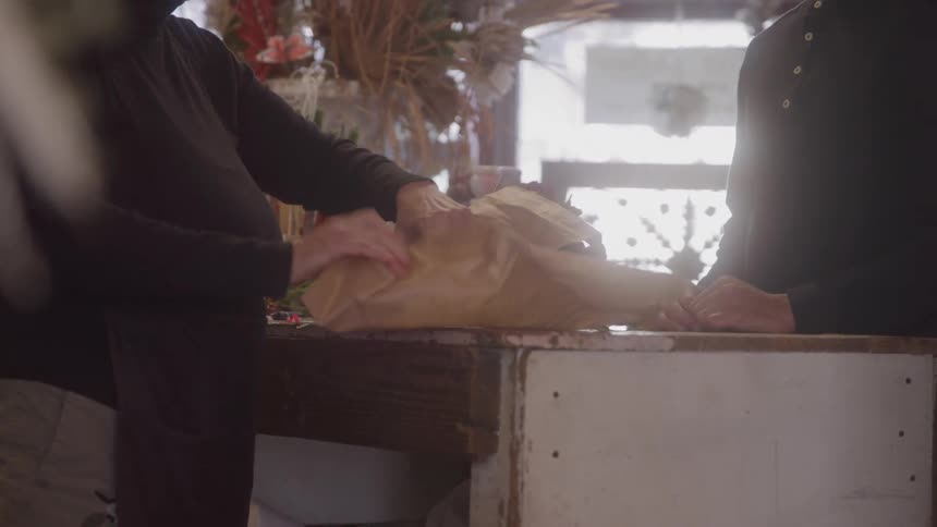 A saleswoman wraps up a flower flower bouquet bought by a man at the counter inside a flower shop at midday