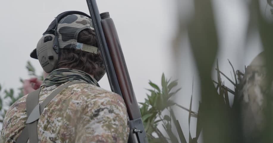 Man in camouflage clothing and ear protection holds a shotgun amid vegetation
