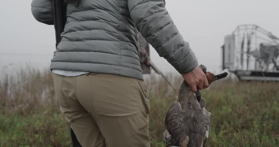 A woman carries a dead bird through a field