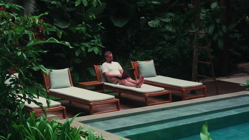 A man reclines on a poolside lounge chair working on a laptop in a lush tropical setting