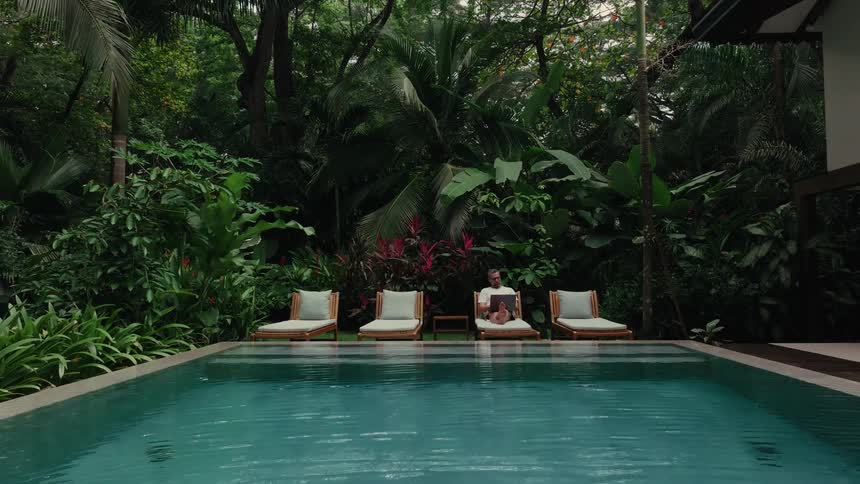 Man sitting on a lounge chair with a laptop on his lap by the poolside surrounded by dense tropical vegetation