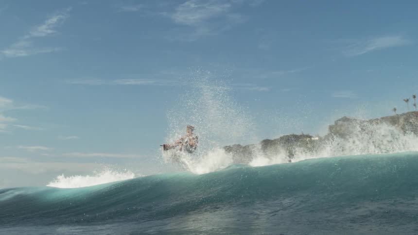 A man surfs on a large turquoise ocean wave catching the white foam as he stands and glides toward the shore which is a rocky cliff with small palm trees on top