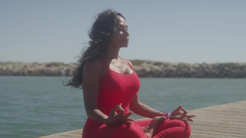 A woman wearing red tights and a dress practices yoga while sitting on a dock at midday