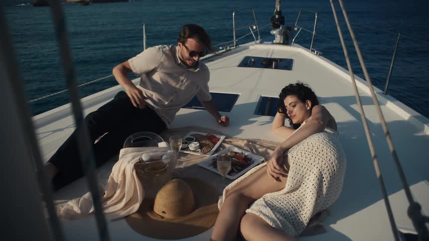 A smiling man and woman are relaxing on the deck of a boat the man is wearing sunglasses and they are sitting on blankets with food and drinks arranged around them
