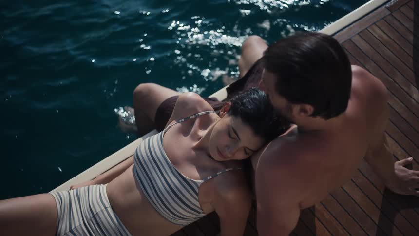 A man and woman are sitting on the deck of a boat with the woman resting her head on the mans shoulder they are both seen from above with the deep blue water of the ocean surrounding them
