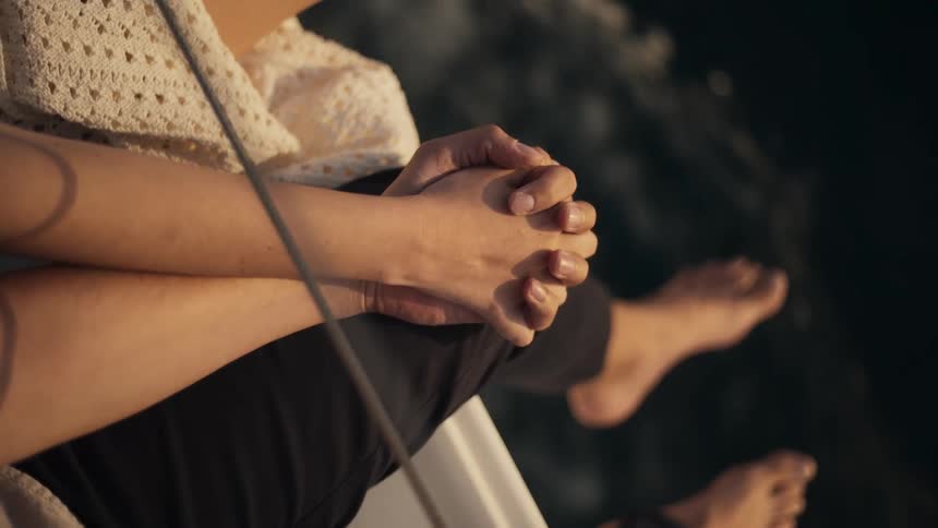 A couple sits close together on a boat holding hands and interlocking their fingers as their bare feet dangle near the water