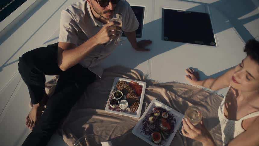 A man in a tan shortsleeved shirt and sunglasses and a woman in a white dress are having a meal on a boat deck sitting on a blanket and raising their wine glasses