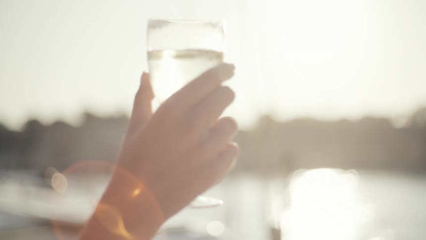 A person takes a flute of champagne on a boat raising the glass and taking a sip while a platter of assorted fruit sits on a blanket nearby